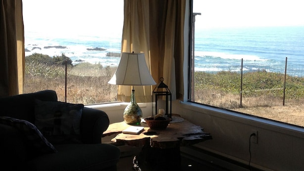 A room with a view of the ocean, a lamp, a candle, and a small table with a book and a bowl. - Cottage by the Sea - Ocean Front (Yachats)
