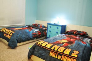 Ein Schlafzimmer mit zwei Betten, jedes mit einer Bettdecke im 'Iron Man 2'-Design. Dazwischen steht ein Nachttisch mit einer Lampe.
