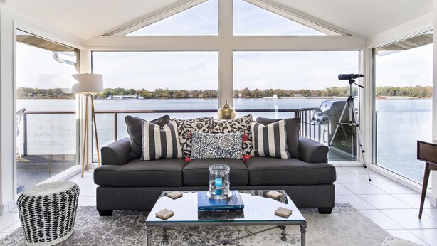 Ein modernes Wohnzimmer mit großem Fenster mit Seeblick, einer Couch mit gemusterten Kissen, einem Glastisch und einer Lampe.