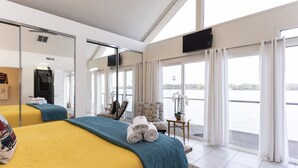 Ein Hotelzimmer mit zwei Betten, einem großen Fenster mit Vorhängen, einem Fernseher und Blick auf das Wasser.