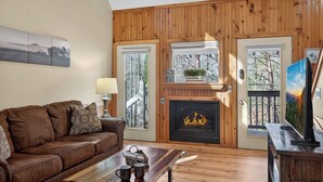 Ein gemütliches Wohnzimmer mit braunem Ledersofa, einem Holztisch, einem Kamin und einem Fernseher auf einem Holzständer.