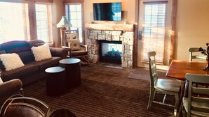 A cozy living room with a stone fireplace, a comfortable sofa, a dining table, and a television mounted on the wall. - Creekside Condo w/ private hot tub at Boyne!  Ski-in/Ski-out, Golf, Bike, Swim  (Boyne Falls)