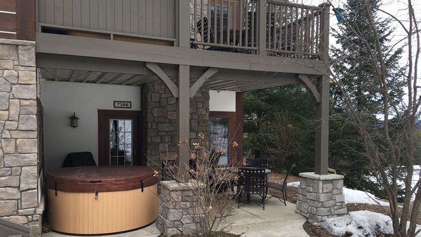 A two-story building with a stone facade, a hot tub, and a covered patio area. - Creekside Condo w/ private hot tub at Boyne!  Ski-in/Ski-out, Golf, Bike, Swim  (Boyne Falls)