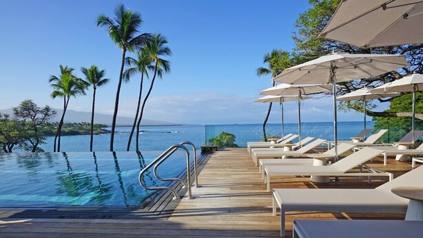 Um deck de madeira com piscina, cadeiras de sol e guarda-sóis, com vista para o mar azul e palmeiras.