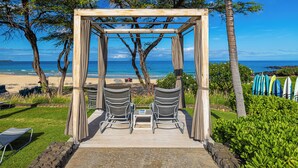 Um gazebo de madeira com duas cadeiras e uma mesa, com vista para uma praia e pranchas de surf ao fundo.