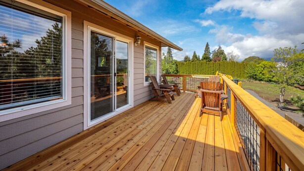 Eine Holzterrasse mit Sitzgelegenheiten im Freien und großen Fenstern mit Blick auf einen Garten.