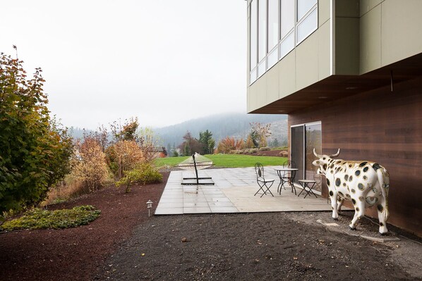 A modern building with a patio area featuring a decorative bull sculpture, outdoor seating, and a view of a mountainous landscape. - Mountain House - Luxury, Rural Setting, Stunning Views (Mount Hood Parkdale)