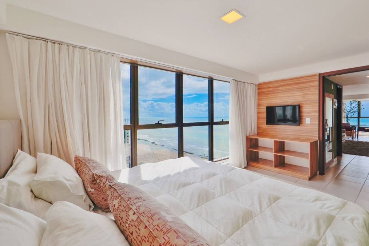 A bedroom with a large bed, a TV mounted on a wooden wall, and a view of the sea through large windows.