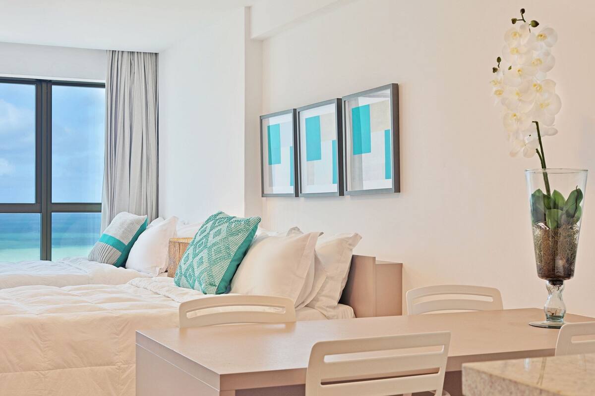 A hotel room with a bed, a dining table, chairs, a vase with flowers, and framed artwork on the wall.
