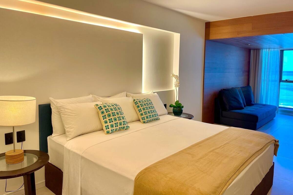 A hotel room with a bed, bedside table, lamp, and a view of the outside.