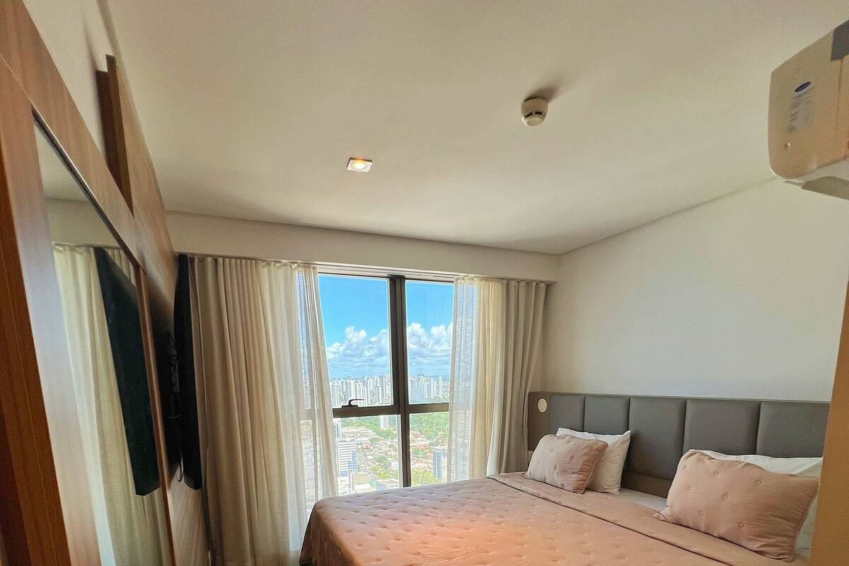 A hotel room with a large bed, a view of the city through a window, and a ceiling-mounted light fixture.