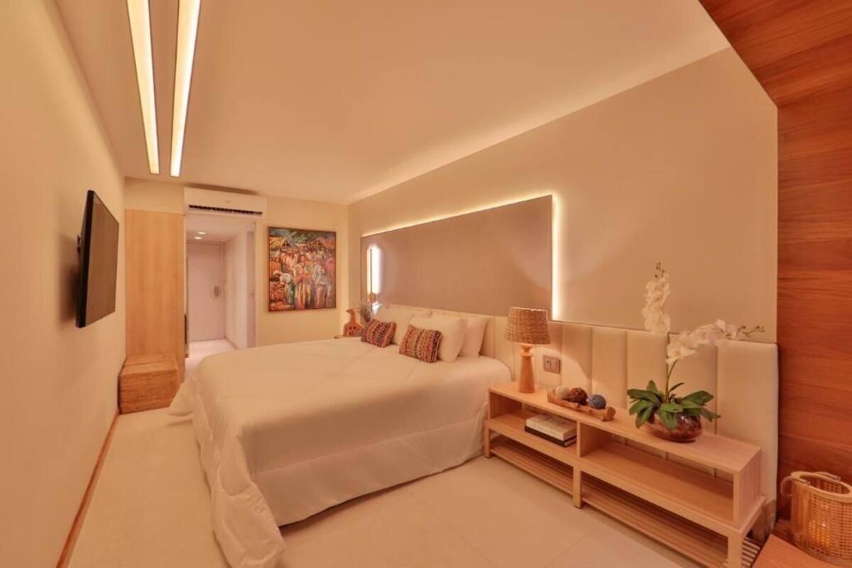 A modern bedroom with a large bed, a bedside table with a lamp, a wall-mounted TV, and a painting on the wall.