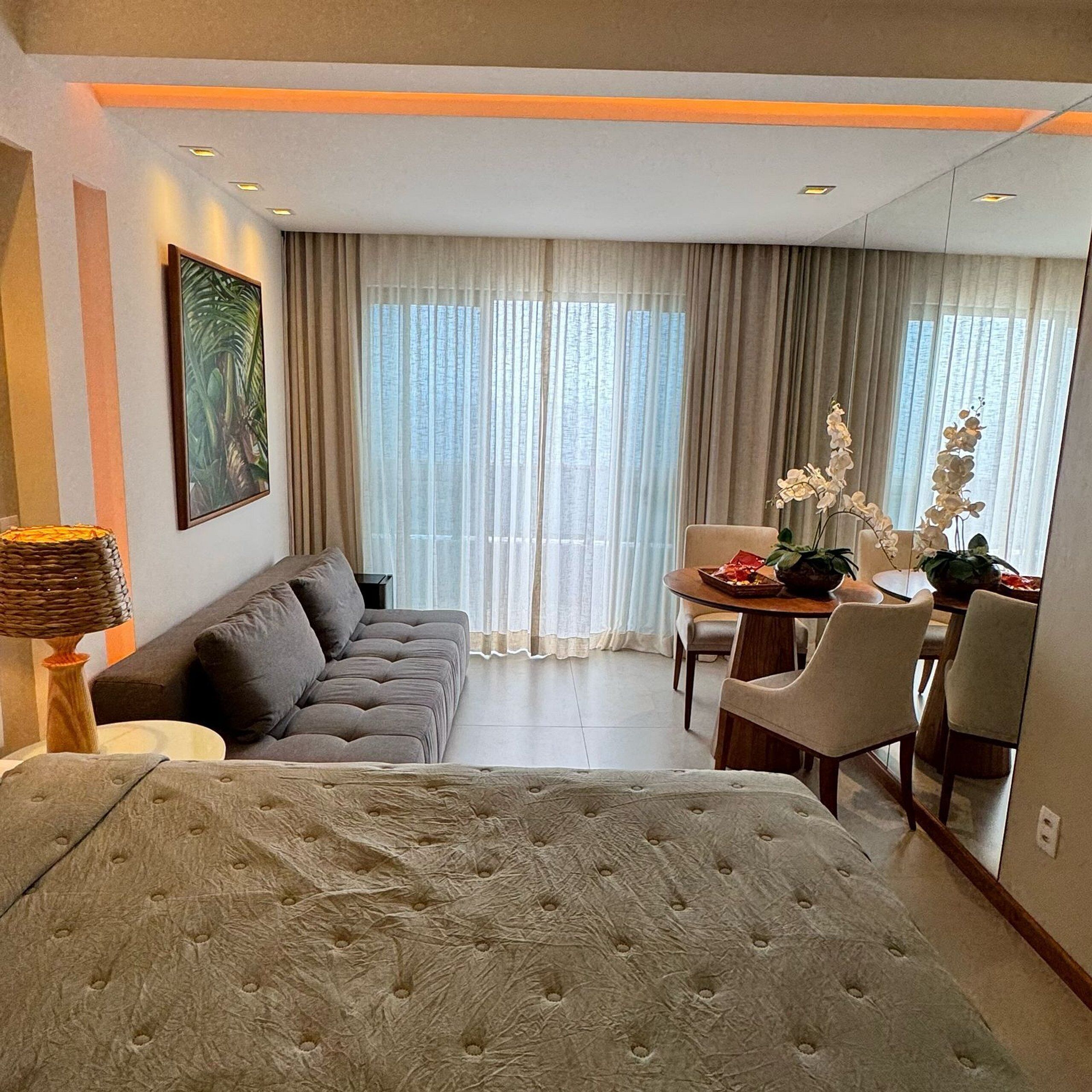 Quarto de hotel moderno com uma cama grande, um sofá, uma mesa de jantar com cadeiras e vista para o exterior através de cortinas leves.