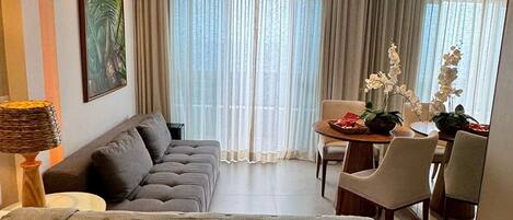 Quarto de hotel moderno com uma cama grande, um sofá, uma mesa de jantar com cadeiras e vista para o exterior através de cortinas leves.