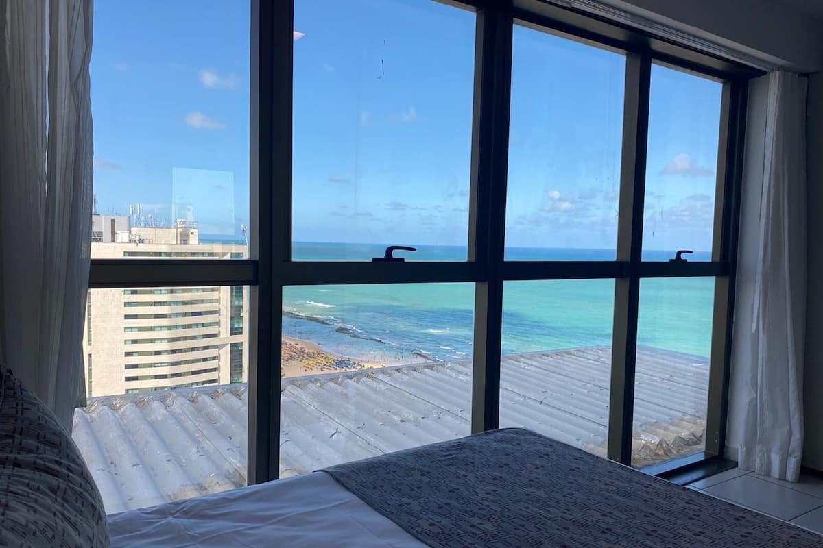 A room with a large window overlooking a beach and ocean, a bed with a patterned bedspread, and a view of a cityscape in the background.