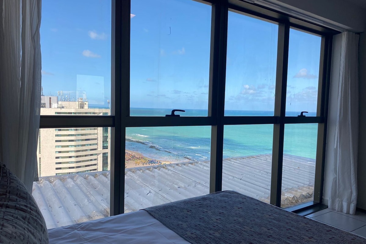 A room with a large window overlooking a beach and ocean, a bed with a patterned bedspread, and a view of a cityscape in the background.