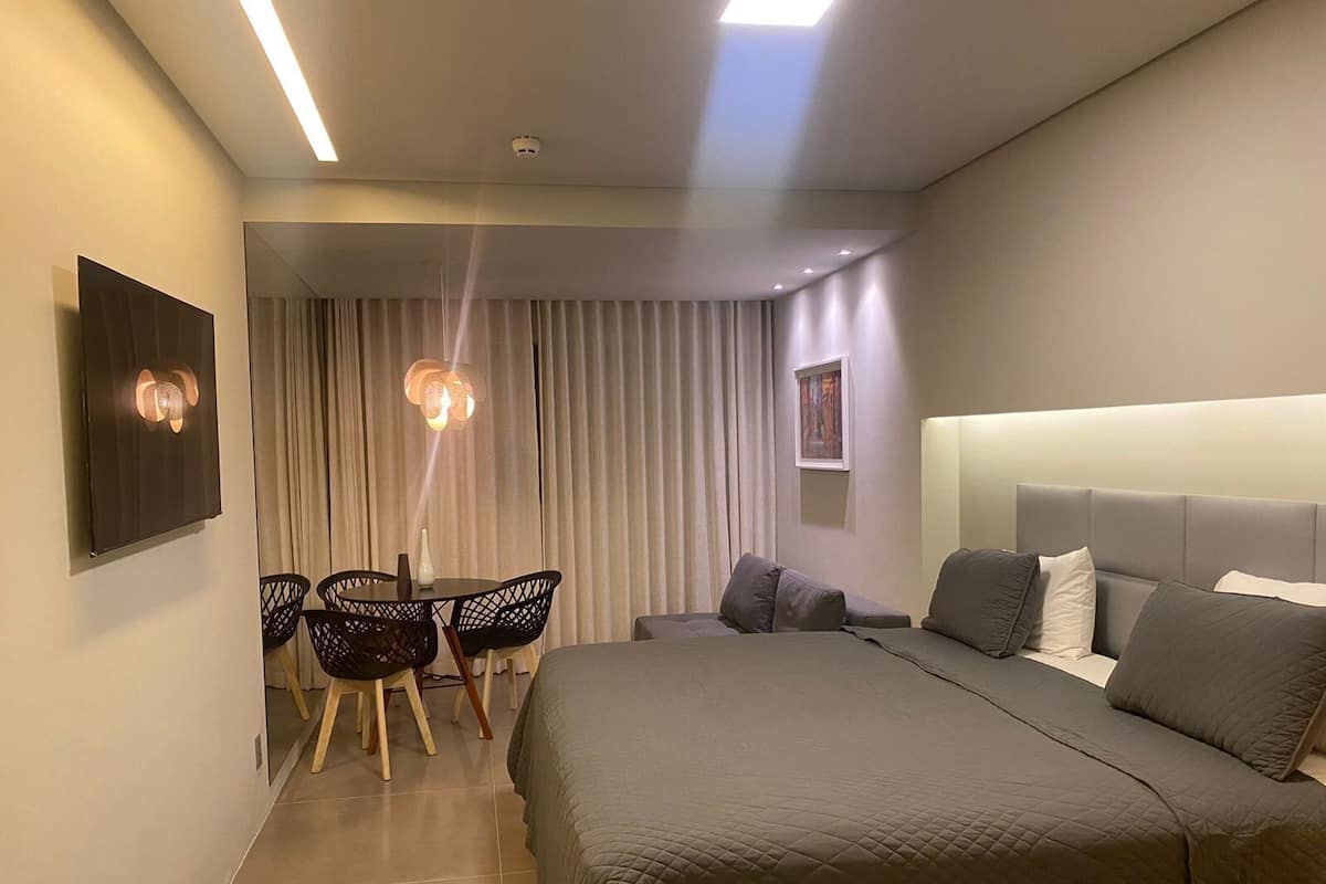 A modern hotel room with a large bed, a dining area with chairs, and a view of the city through the window.
