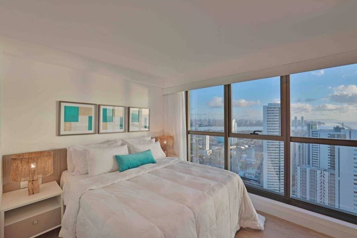 A modern bedroom with a large window, a bed with white bedding, a nightstand, and framed artwork on the wall.
