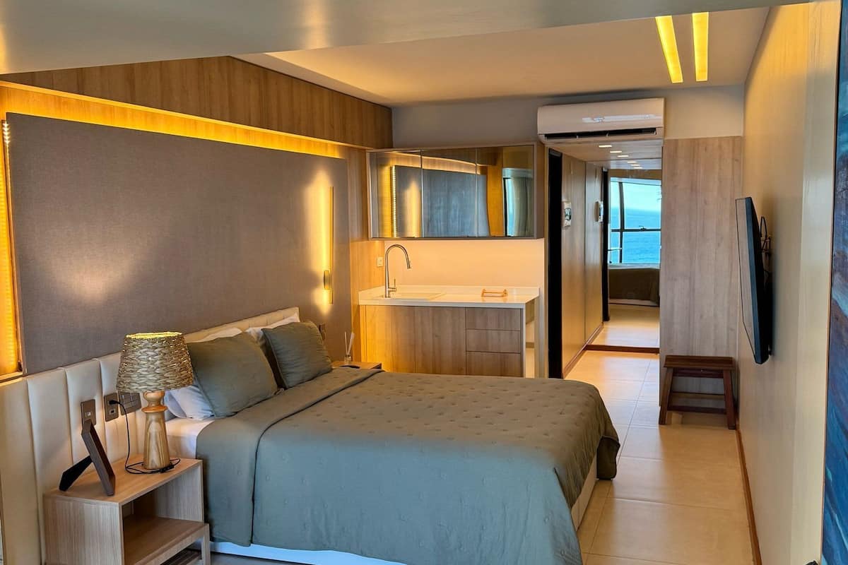 Una habitación de hotel moderna con una cama grande, mesita de noche, lámpara y vista al mar a través de una ventana.