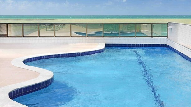 A swimming pool with a curved edge, a glass railing, and a view of the beach and ocean. - HY Beach Flats (Recife)