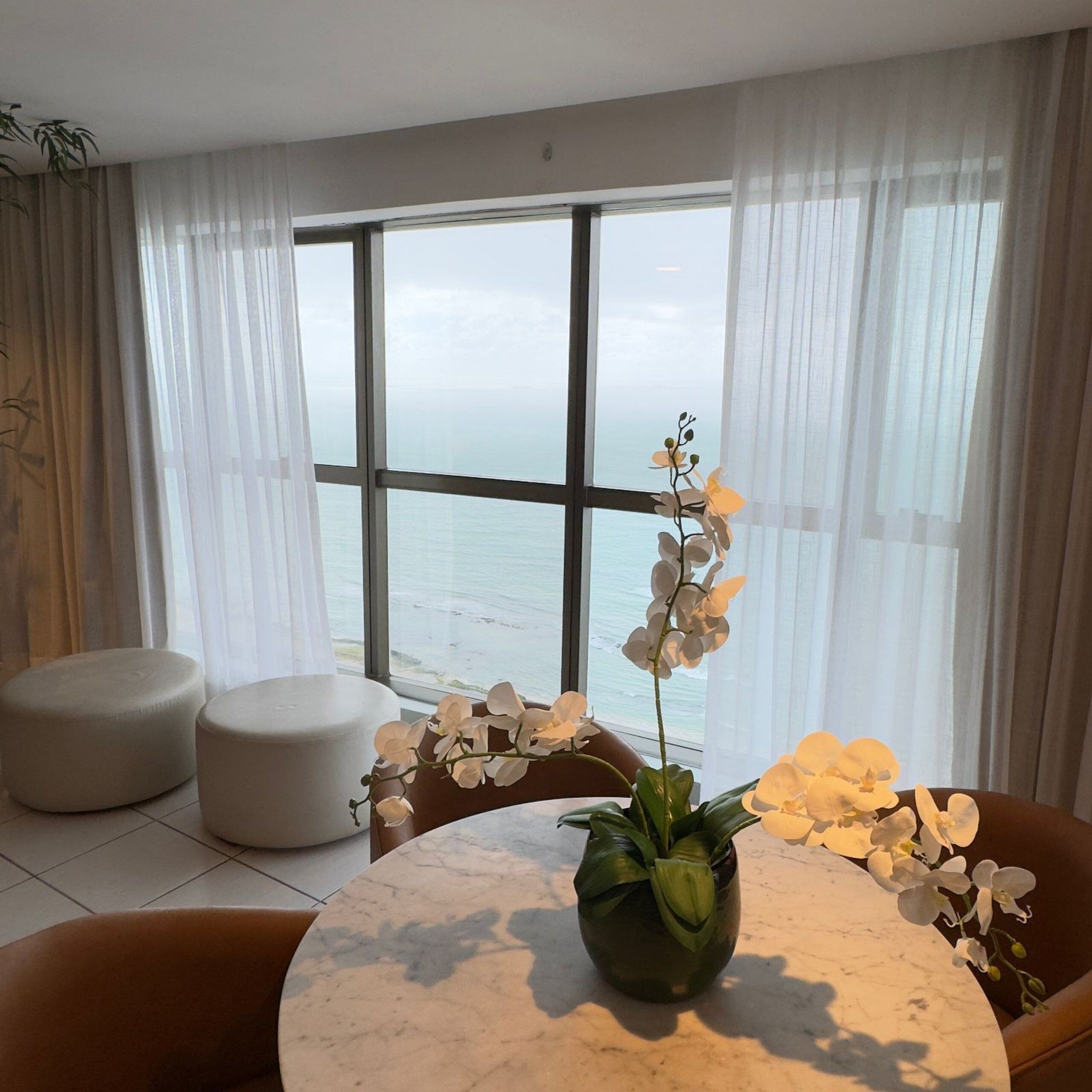 A modern living room with a large window overlooking the sea, a round marble table, and two round ottomans.