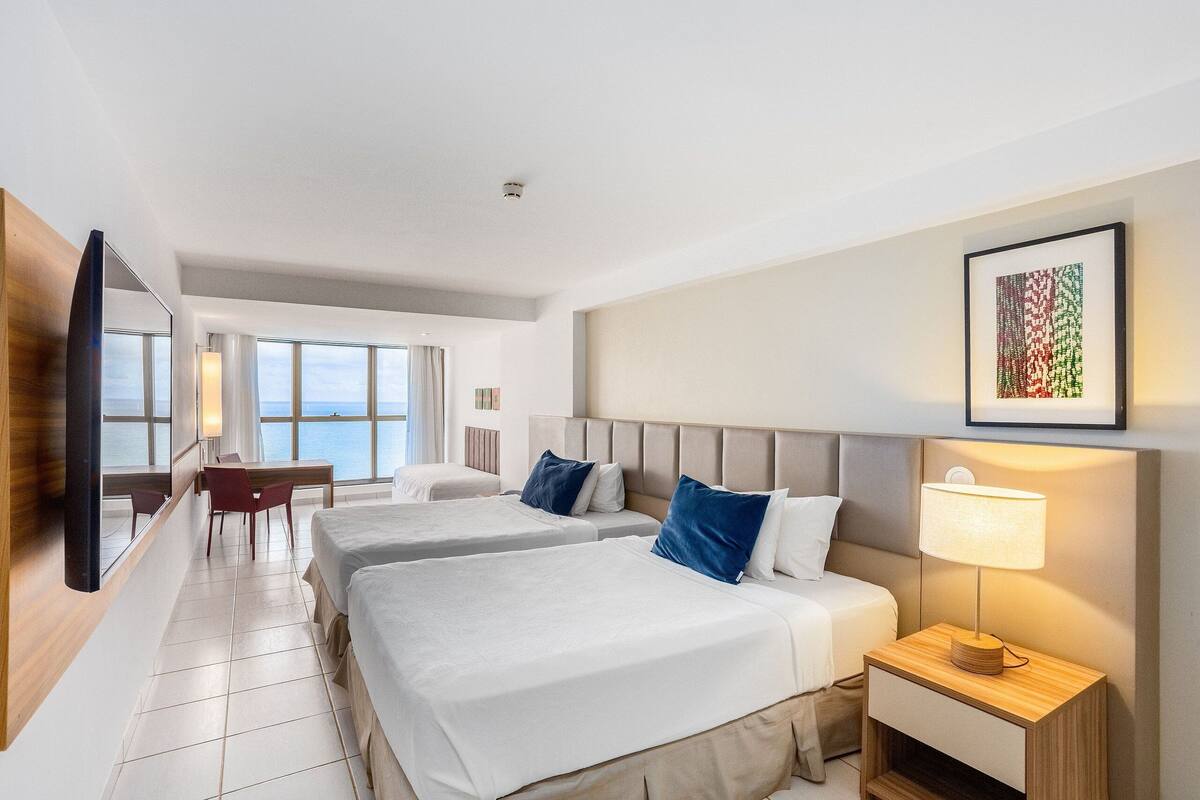 A hotel room with two beds, a desk, and a view of the sea.