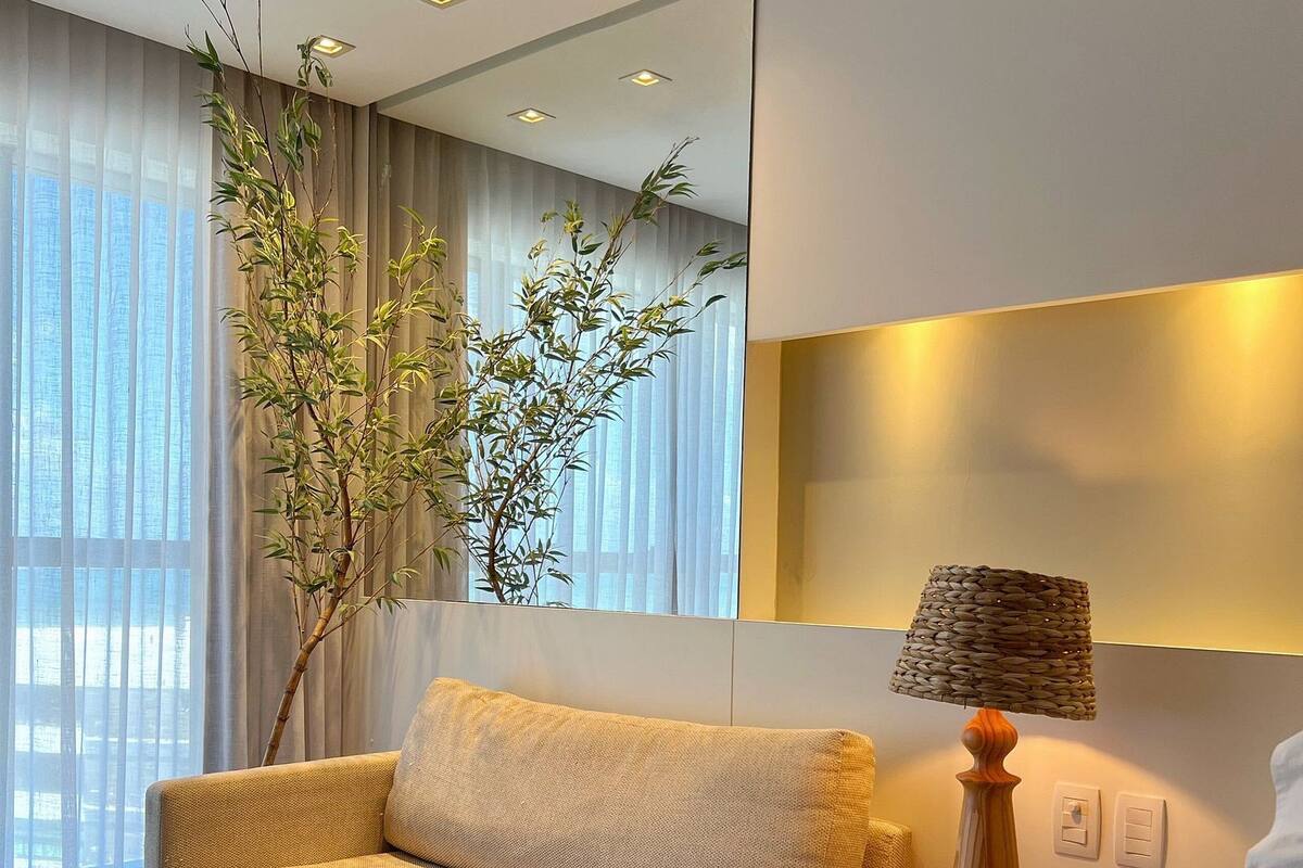 A modern hotel room with a sofa, a bed, a bedside table with a lamp, and a large window with sheer curtains.