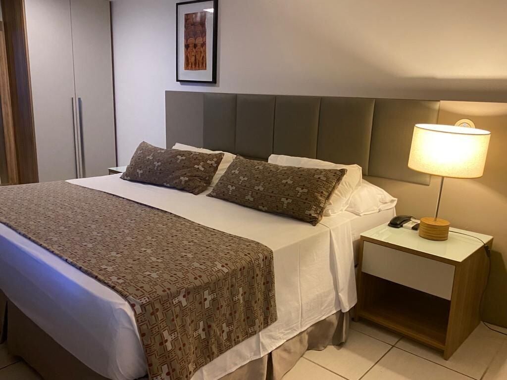 Una cama bien hecha con ropa de cama estampada, una lámpara de noche y una mesita de noche pequeña con un teléfono y una lámpara.