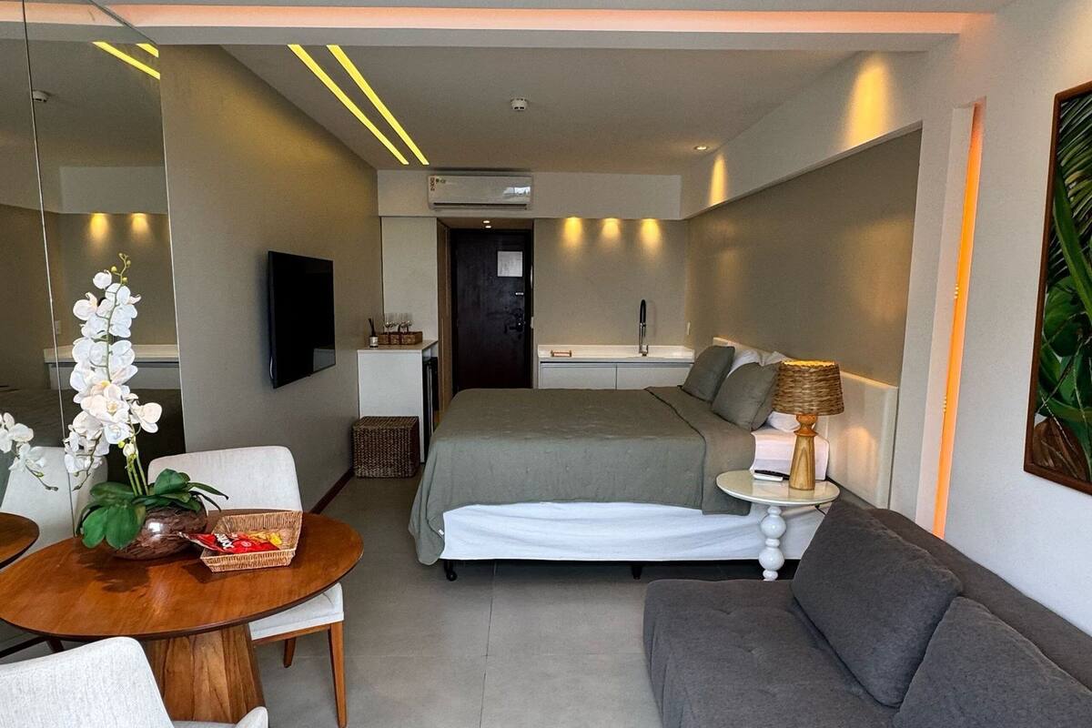 Habitación de hotel moderna con cama, sofá, zona de comedor con mesa y sillas, televisor y una pequeña kitchenette.