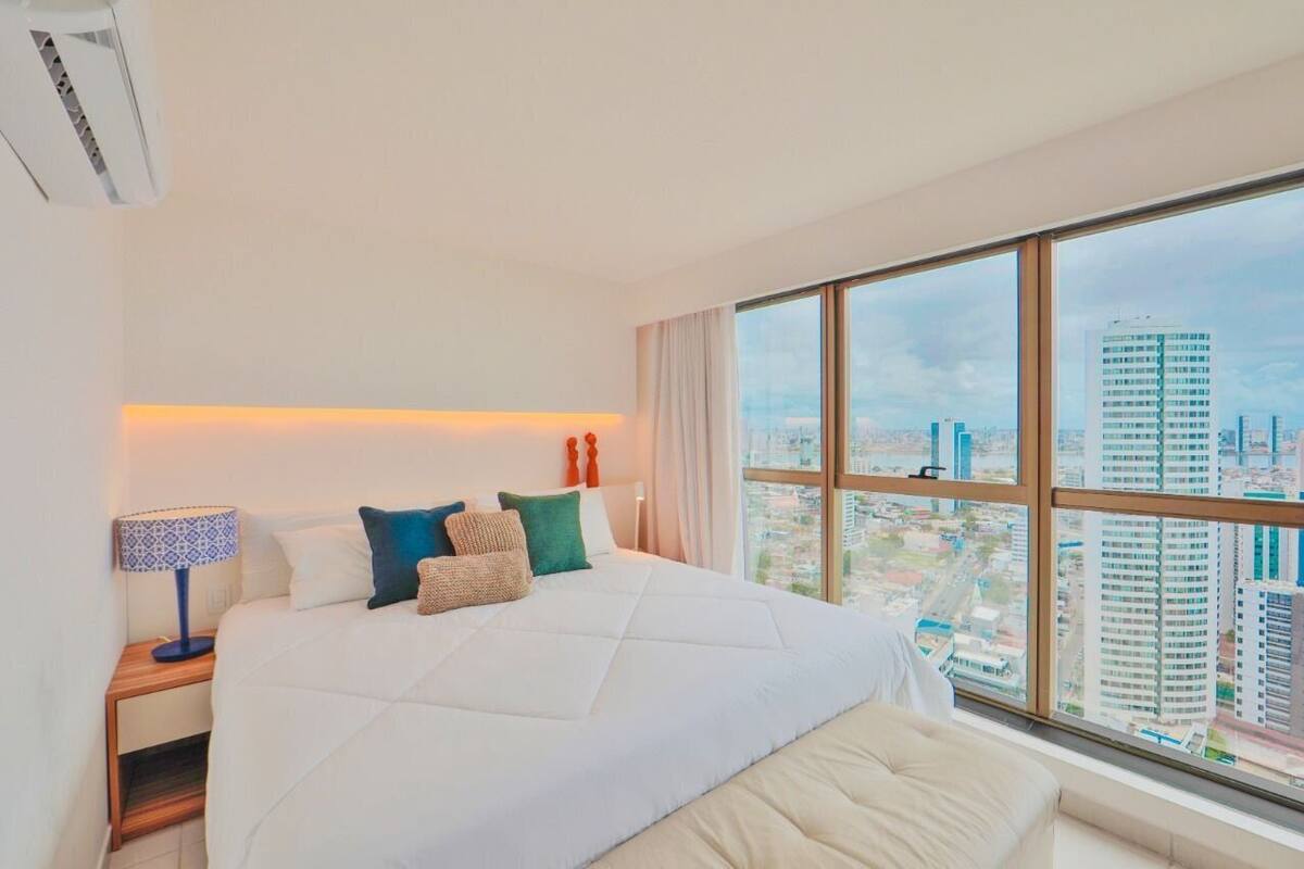 A hotel room with a large bed, a nightstand with a lamp, and a view of the city through large windows.