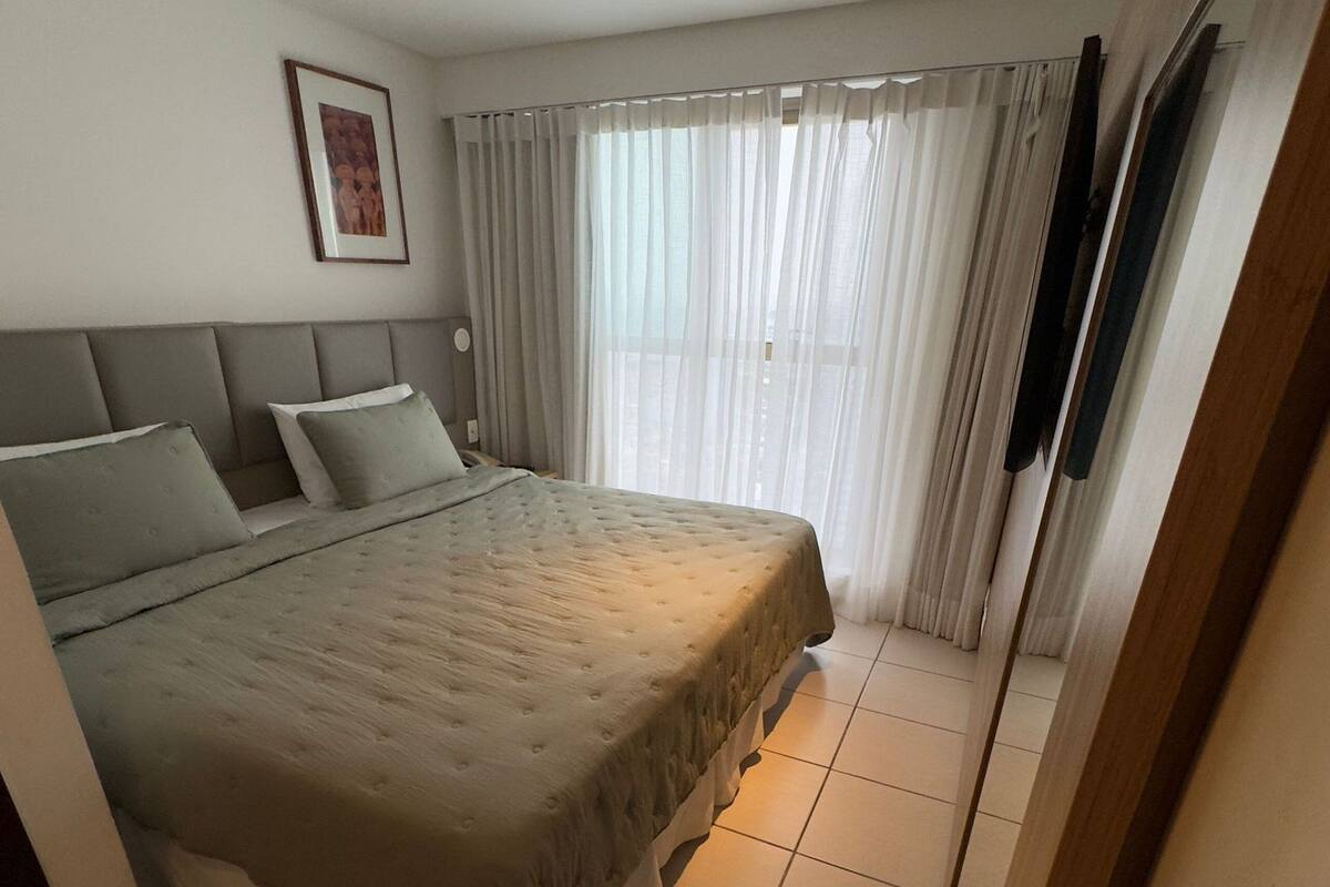 A bedroom with a large bed, a headboard, a framed picture on the wall, and a window with curtains.