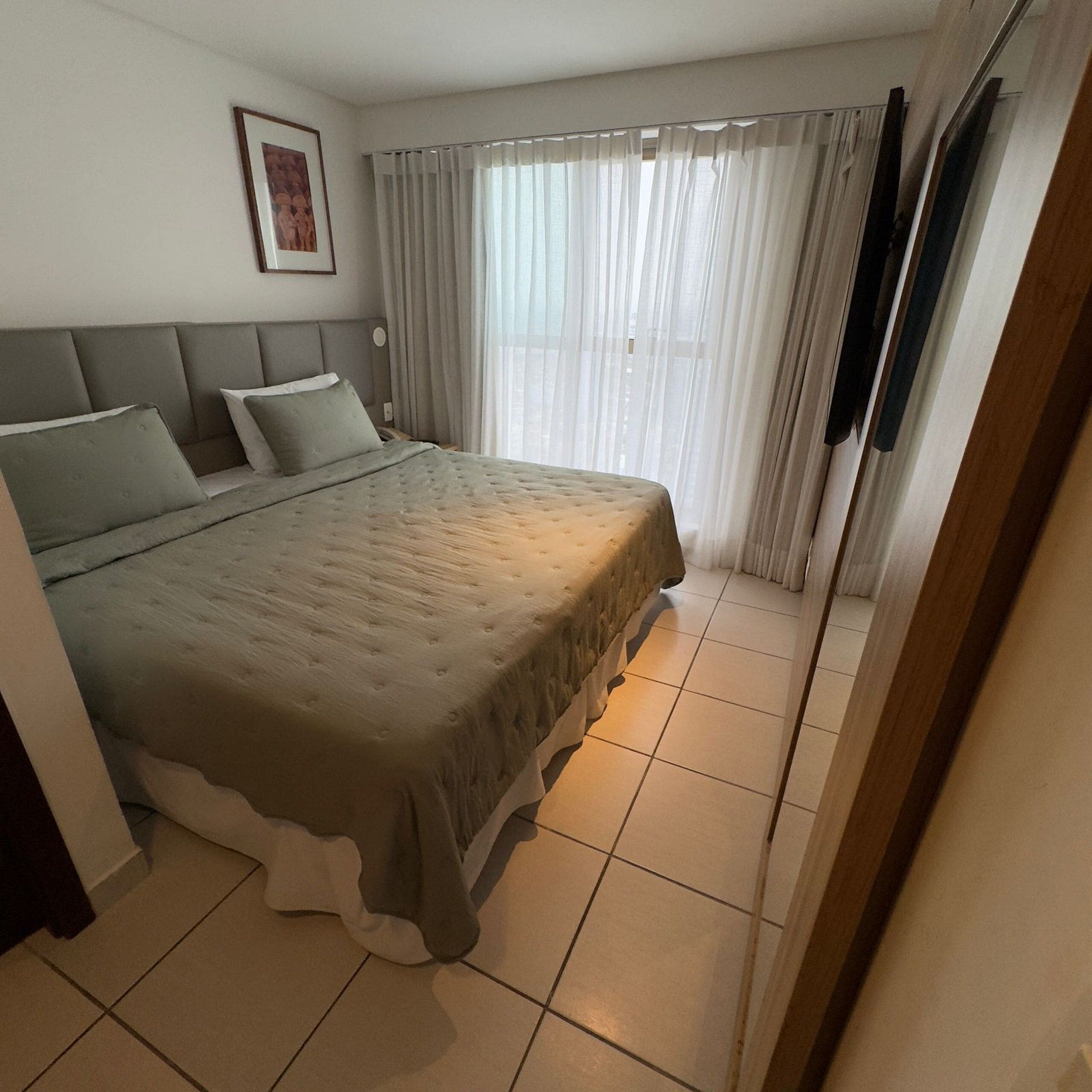 A bedroom with a large bed, a headboard, a framed picture on the wall, and a window with curtains.