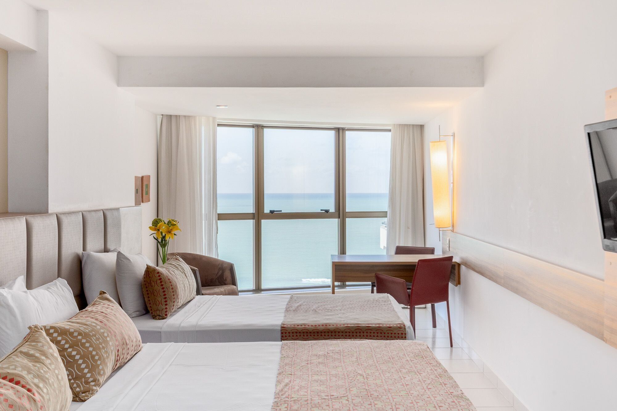 A hotel room with two beds, a desk, a chair, a TV, and a large window with a view of the sea.