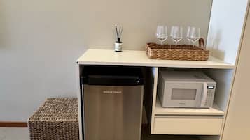 Una kitchenette compacta con un refrigerador de acero inoxidable, un microondas y una cesta tejida sobre un estante blanco.
