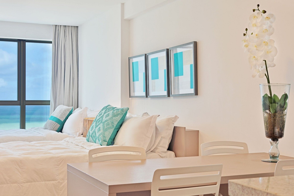 A hotel room with a bed, a dining table, chairs, a vase with flowers, and framed artwork on the wall.