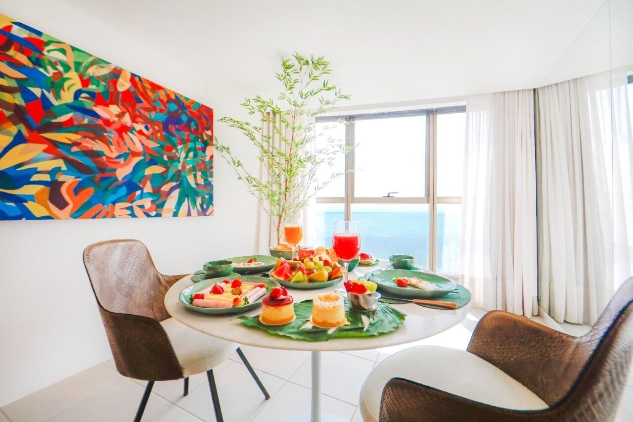 Un coin repas avec une table ronde dressée pour deux personnes, entourée de chaises. On y trouve une grande fenêtre avec des rideaux, une plante en pot et un tableau abstrait aux couleurs vives accroché au mur.