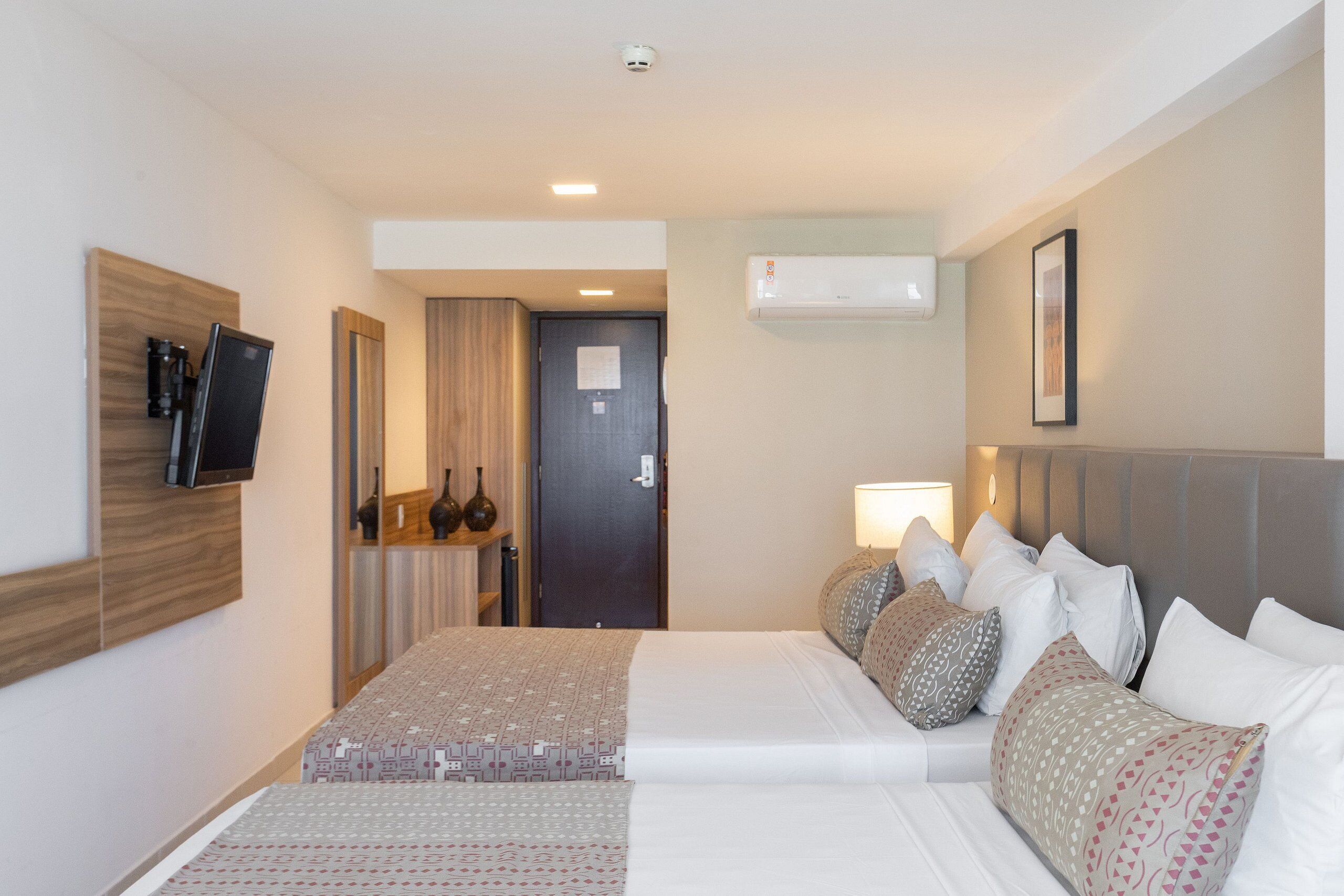 A hotel room with a bed, bedside table, TV, and an air conditioning unit.