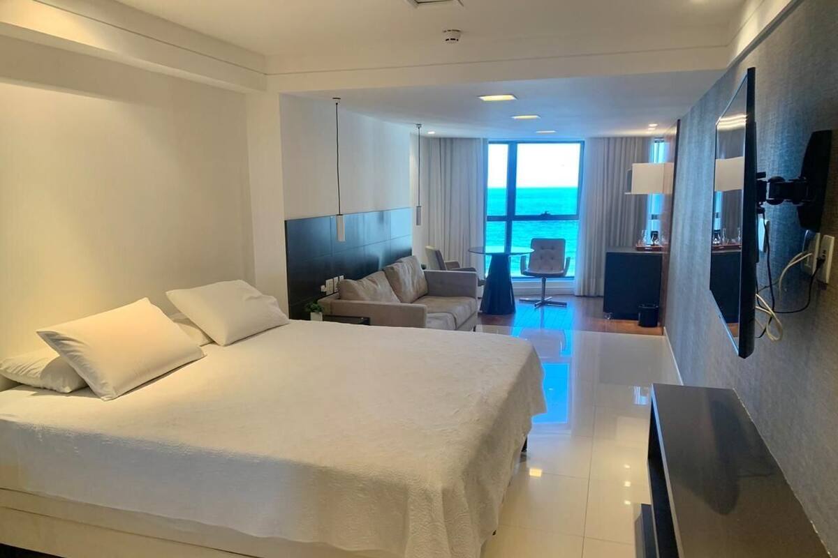 A hotel room with a large bed, a seating area with a sofa and chairs, a flat-screen TV, and a view of the sea through the window.