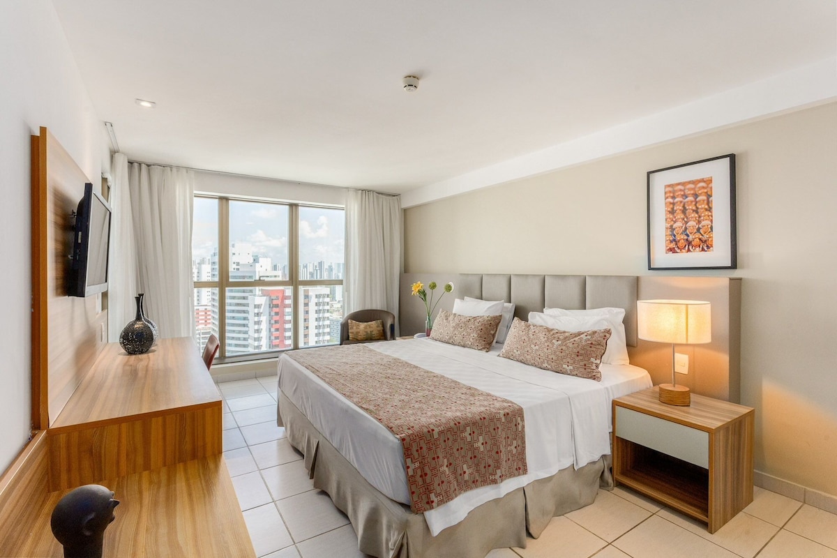 A hotel room with a bed, bedside table, lamp, and a city view through the window.