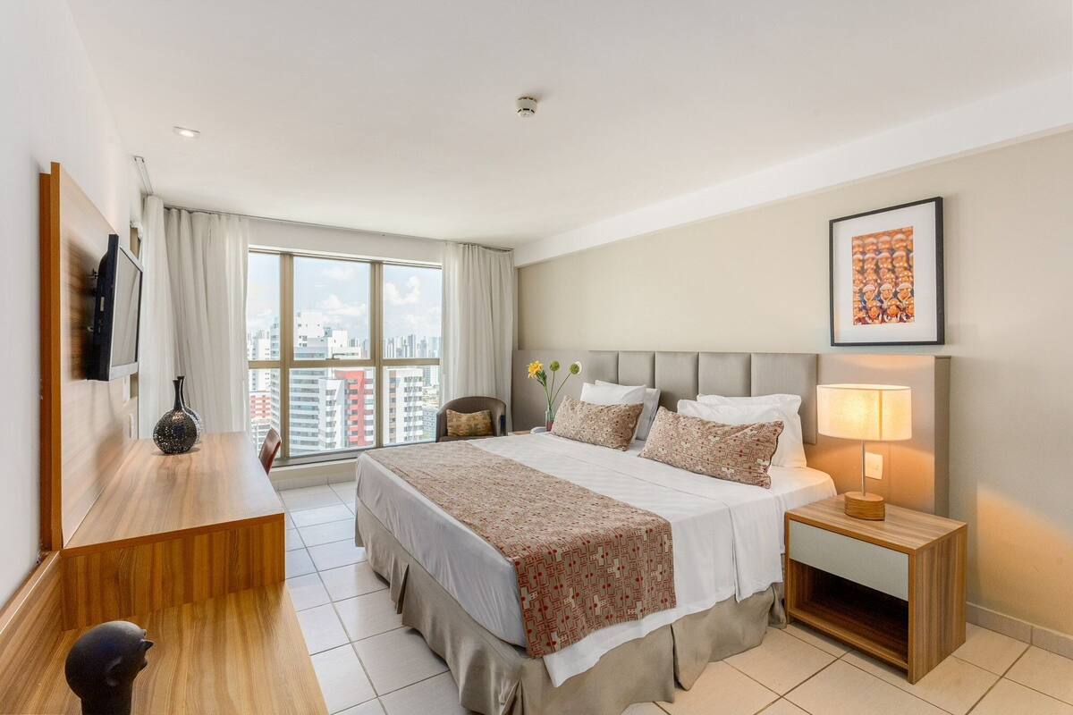 A hotel room with a bed, bedside table, lamp, and a city view through the window.