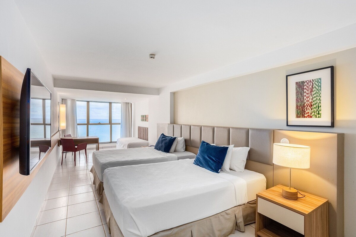 A hotel room with two beds, a desk, and a view of the sea.