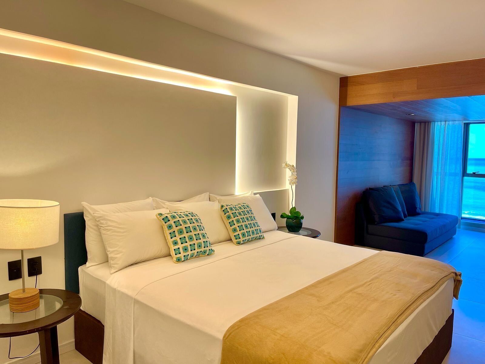 Habitación de hotel con cama, mesita de noche, lámpara y vistas al exterior.