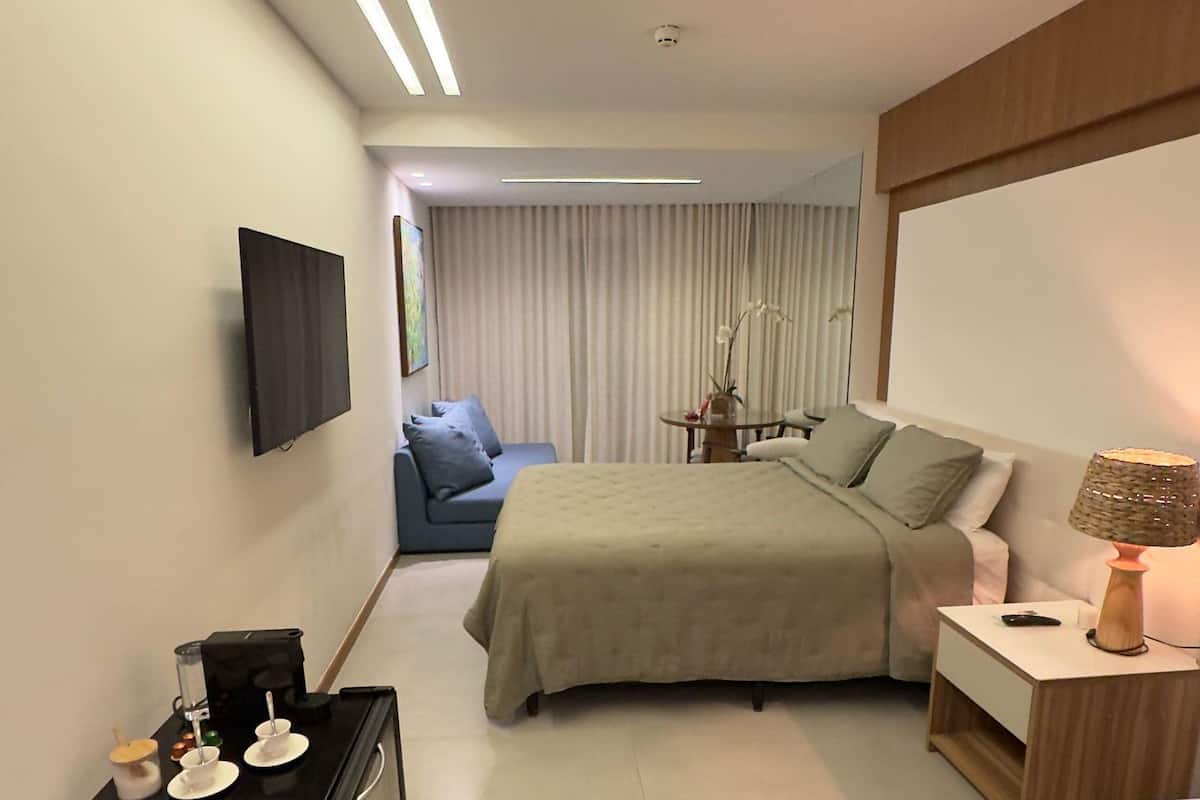 A modern hotel room with a bed, a sofa, a desk with a lamp, and a TV on the wall.