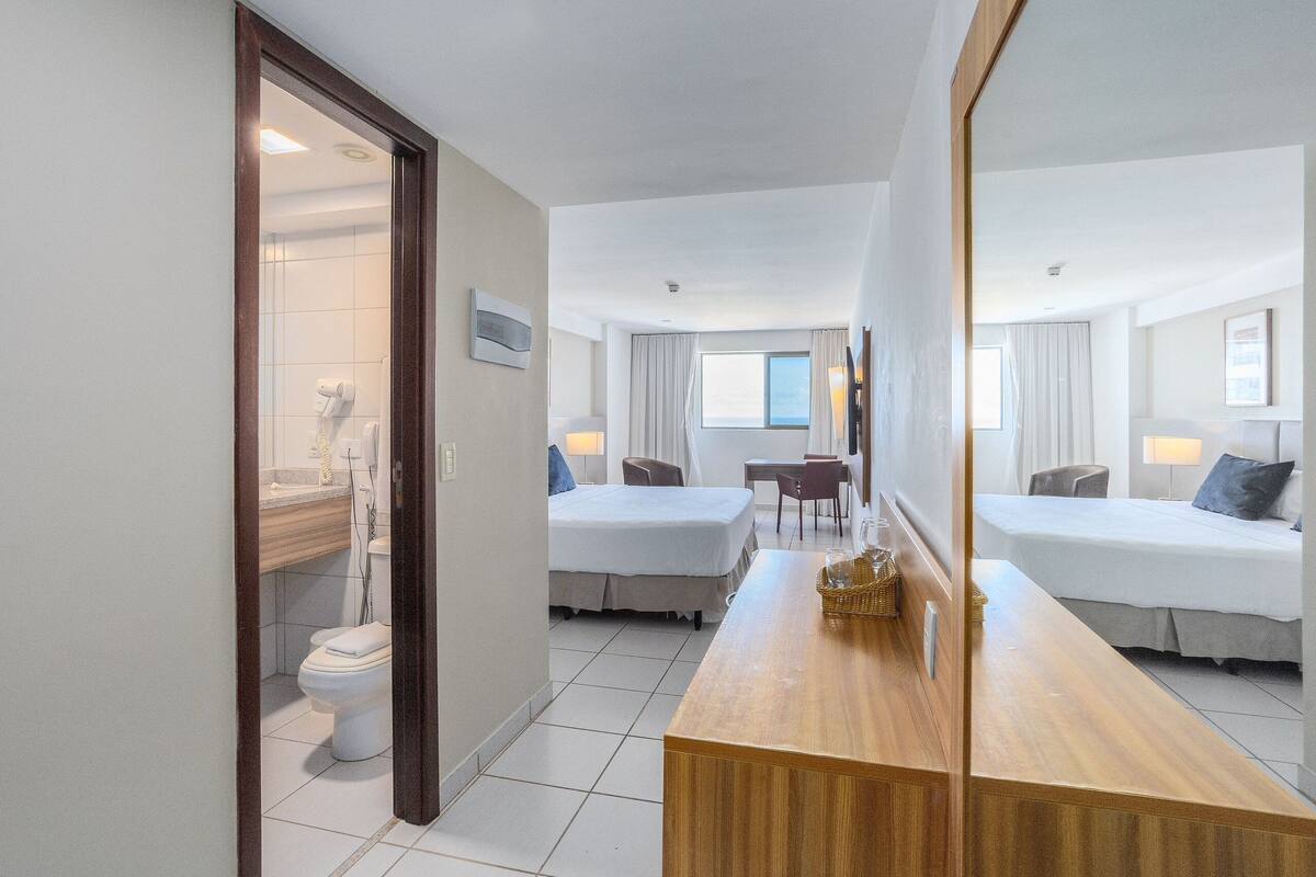 A hotel room with two beds, a wooden desk, a bathroom with a toilet and sink, and a large mirror.