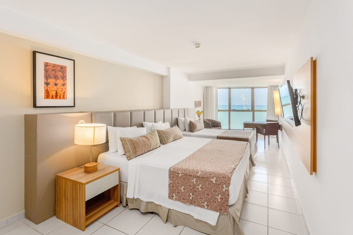 A hotel room with two beds, a desk, a chair, a TV, and a large window with a view of the sea.