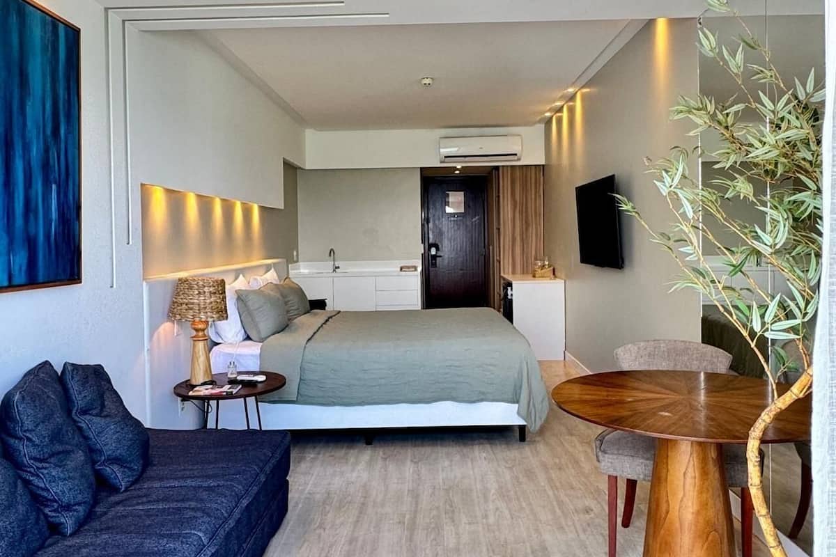 A modern hotel room with a bed, a sofa, a dining table, and a TV.