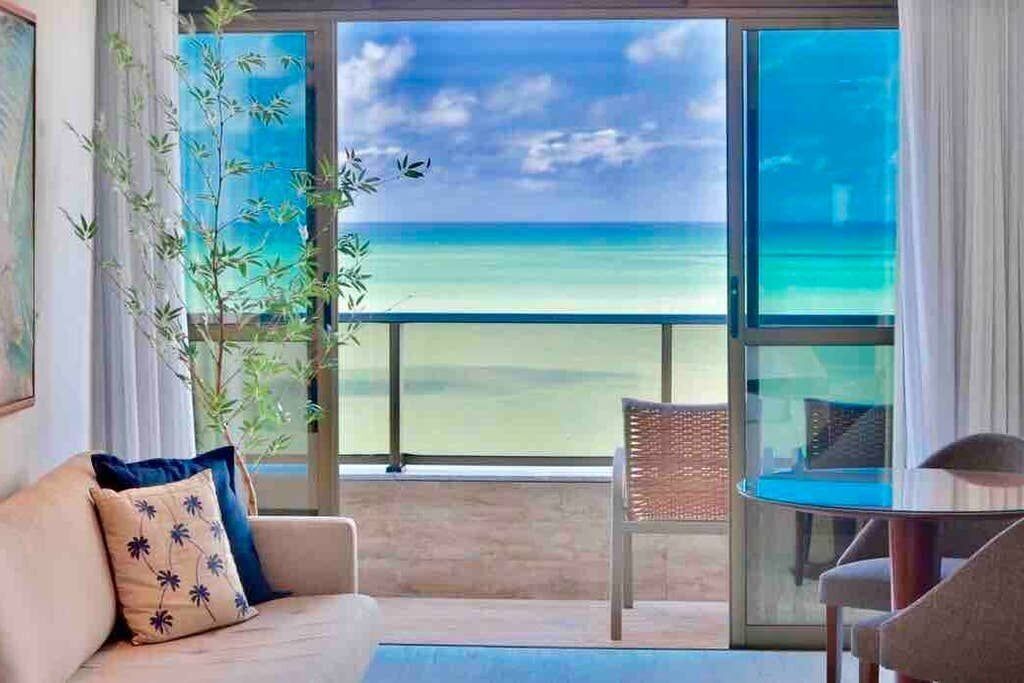 A room with a view of the ocean, a sofa with a decorative pillow, a chair, and a glass table.