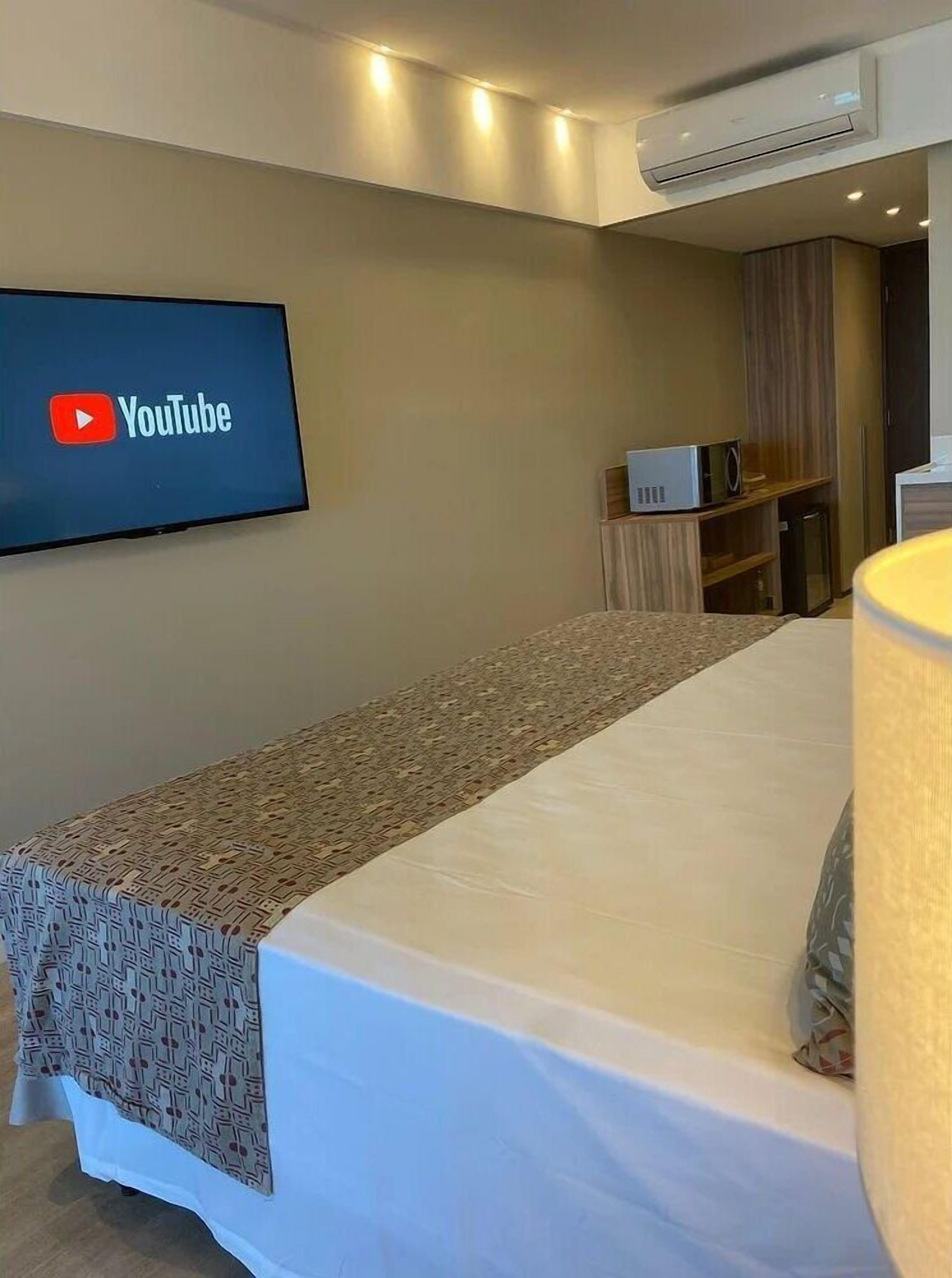 Habitación de hotel con cama, televisión de pantalla plana en la pared mostrando YouTube, unidad de aire acondicionado, microondas y un mueble pequeño.