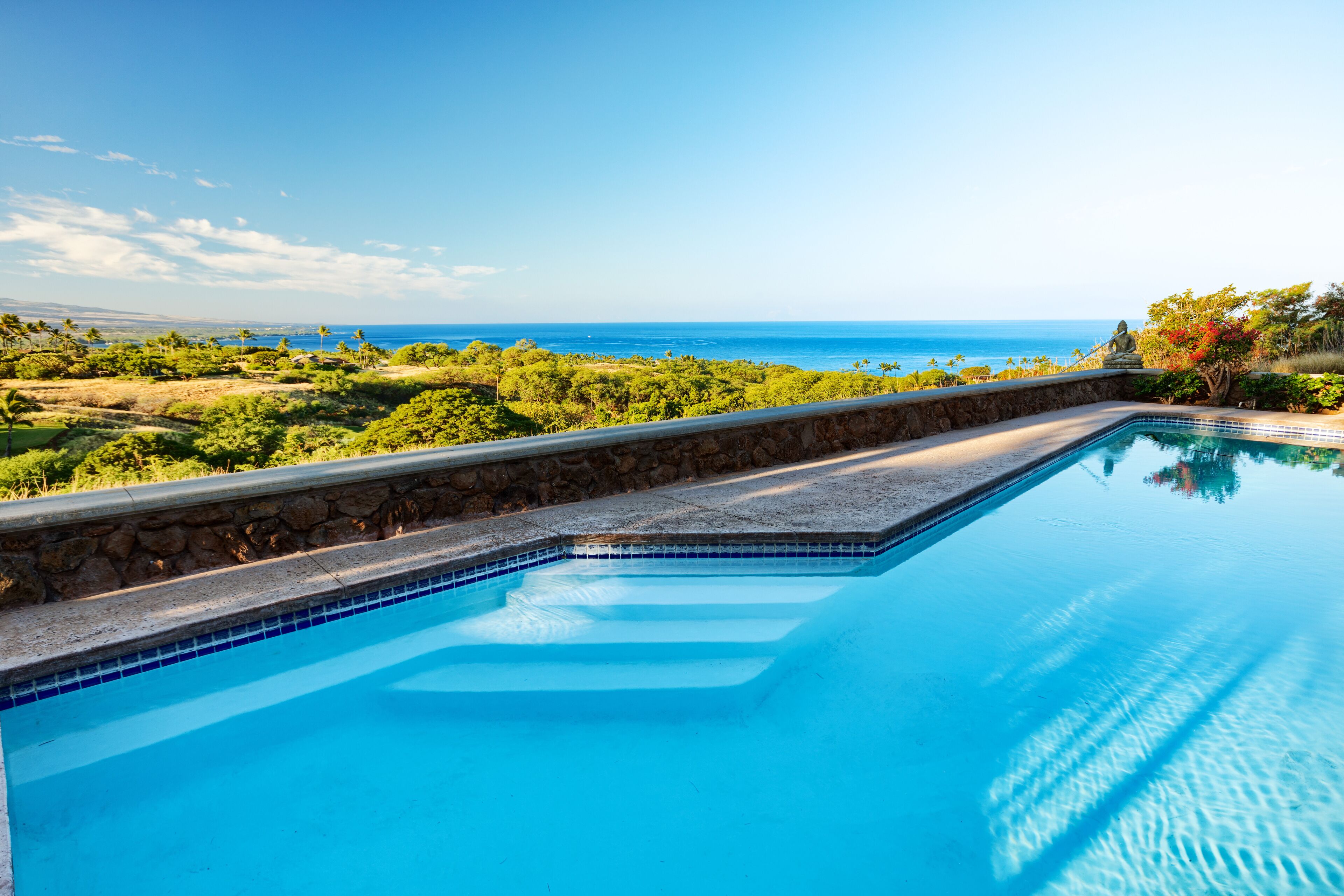 A clear view of a swimming pool with a stone edge and a stone wall, overlooking a lush landscape and the ocean.
