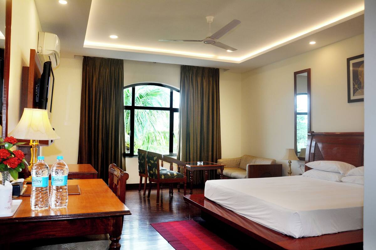 Quarto de hotel com uma cama, uma escrivaninha com candeeiro, uma cadeira, uma mesa e uma janela com cortinas.